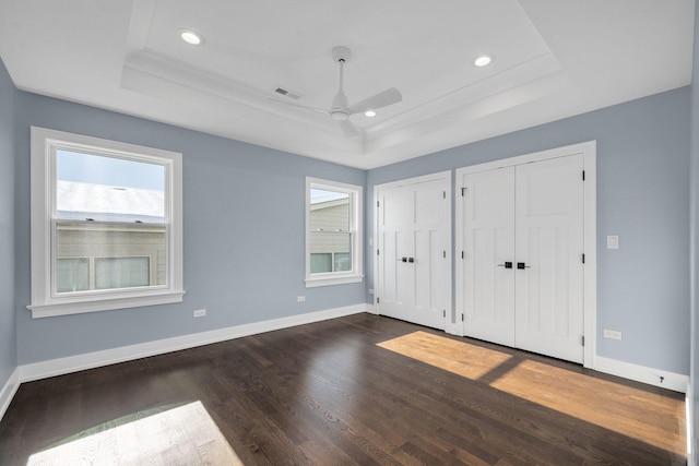 unfurnished bedroom with ceiling fan, a tray ceiling, dark hardwood / wood-style flooring, and multiple closets