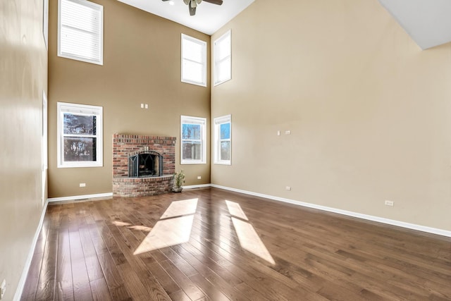 unfurnished living room featuring a towering ceiling, dark hardwood / wood-style flooring, a brick fireplace, and ceiling fan