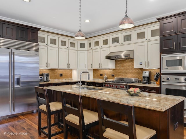 kitchen featuring a kitchen bar, a center island with sink, decorative light fixtures, built in appliances, and wall chimney exhaust hood