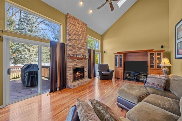 living room with a skylight, ceiling fan, a brick fireplace, high vaulted ceiling, and light hardwood / wood-style flooring