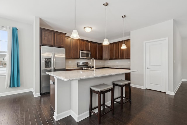 kitchen with light stone countertops, dark hardwood / wood-style flooring, stainless steel appliances, an island with sink, and hanging light fixtures