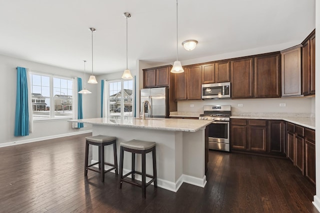 kitchen featuring an island with sink, appliances with stainless steel finishes, dark hardwood / wood-style flooring, hanging light fixtures, and light stone countertops