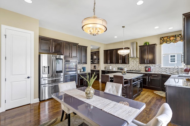 kitchen with appliances with stainless steel finishes, a center island, pendant lighting, light stone counters, and wall chimney range hood