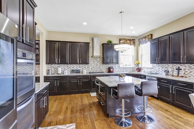 kitchen with stainless steel appliances, wall chimney exhaust hood, hanging light fixtures, light stone countertops, and a kitchen island