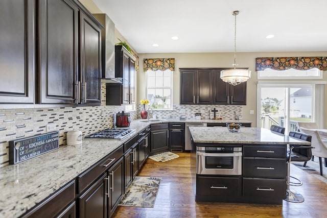 kitchen with wall chimney exhaust hood, hanging light fixtures, stainless steel appliances, tasteful backsplash, and dark brown cabinets