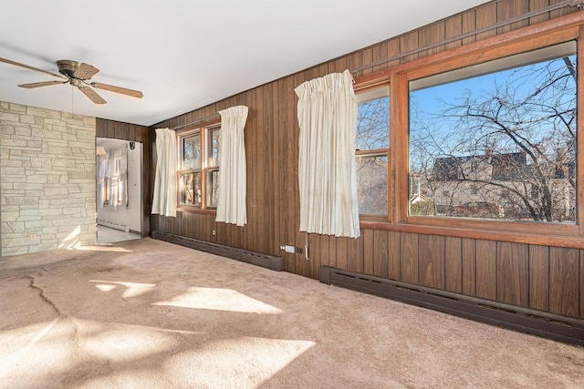 carpeted empty room with a healthy amount of sunlight, a baseboard heating unit, ceiling fan, and wood walls