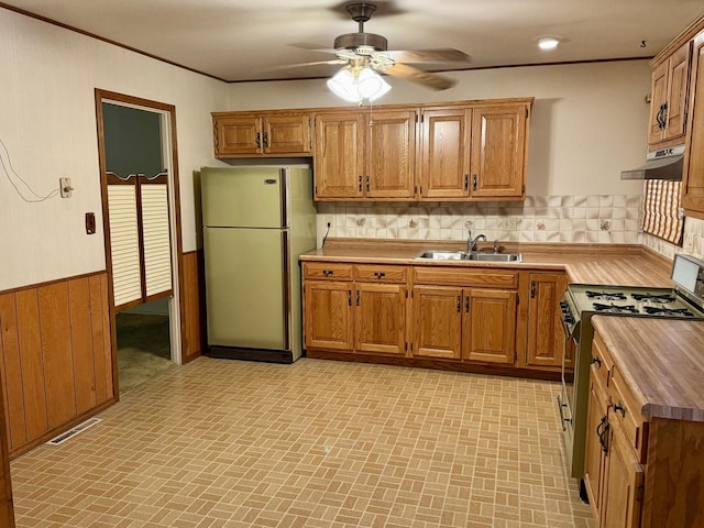 kitchen featuring sink, wood walls, refrigerator, stainless steel gas range oven, and ceiling fan