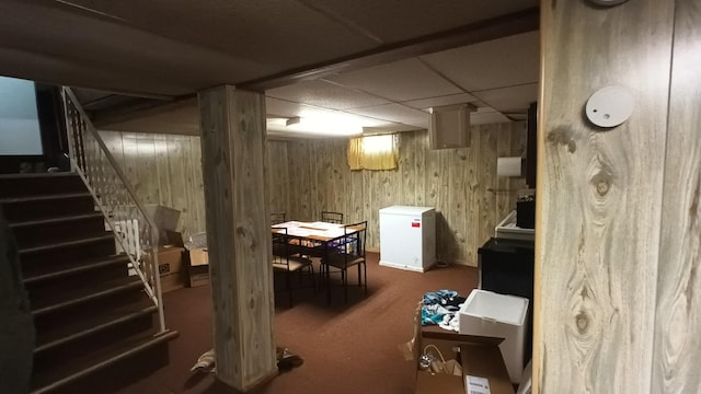 basement featuring wood walls, a drop ceiling, and refrigerator