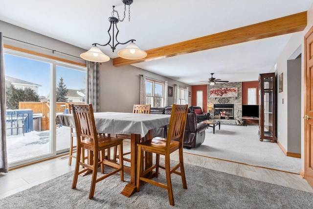 carpeted dining area with a wealth of natural light, ceiling fan, beamed ceiling, and a stone fireplace