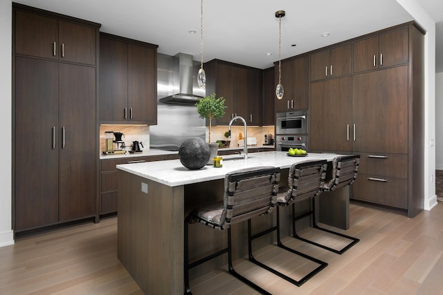 kitchen featuring wall chimney range hood, a kitchen island with sink, decorative light fixtures, and dark brown cabinetry