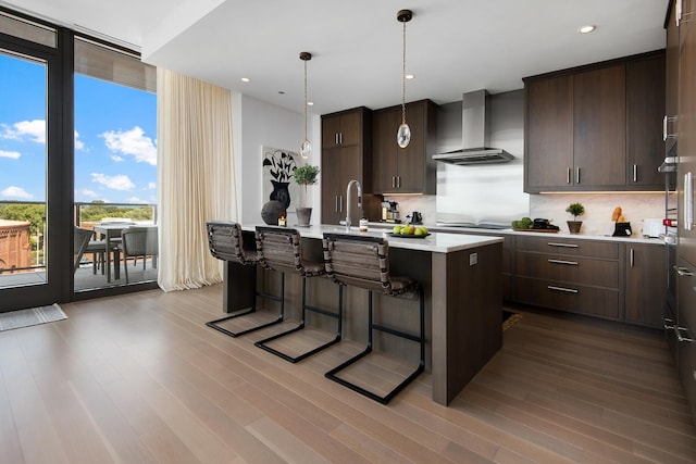 kitchen with a breakfast bar area, a center island with sink, hanging light fixtures, dark brown cabinets, and wall chimney range hood