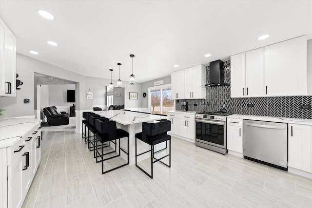 kitchen featuring wall chimney exhaust hood, white cabinetry, decorative light fixtures, appliances with stainless steel finishes, and a kitchen breakfast bar