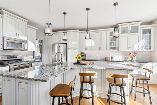 kitchen featuring appliances with stainless steel finishes, an island with sink, sink, hanging light fixtures, and light hardwood / wood-style flooring