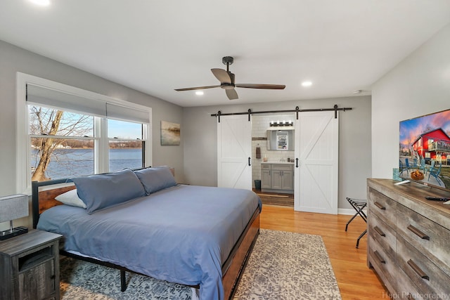 bedroom featuring a barn door, ceiling fan, ensuite bath, a water view, and light hardwood / wood-style flooring