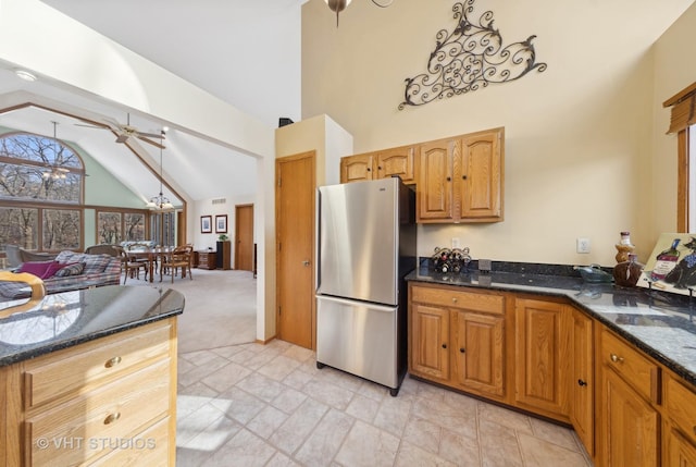 kitchen with high vaulted ceiling, dark stone countertops, stainless steel refrigerator, light colored carpet, and ceiling fan with notable chandelier