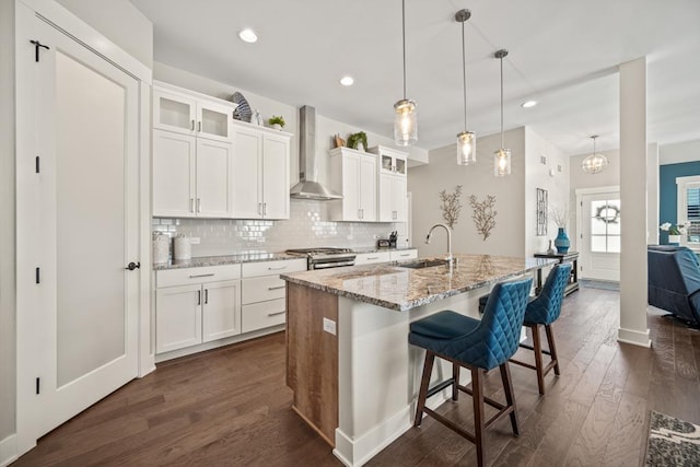 kitchen with white cabinetry, light stone counters, wall chimney exhaust hood, and an island with sink