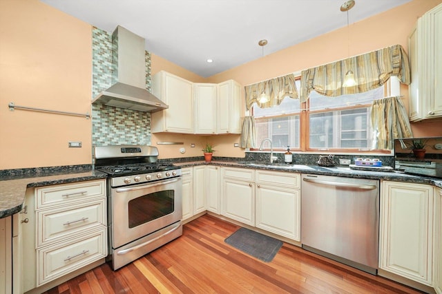 kitchen featuring stainless steel appliances, wall chimney exhaust hood, sink, cream cabinets, and decorative light fixtures