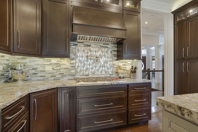 kitchen featuring tasteful backsplash, premium range hood, decorative columns, stainless steel gas stovetop, and dark brown cabinets