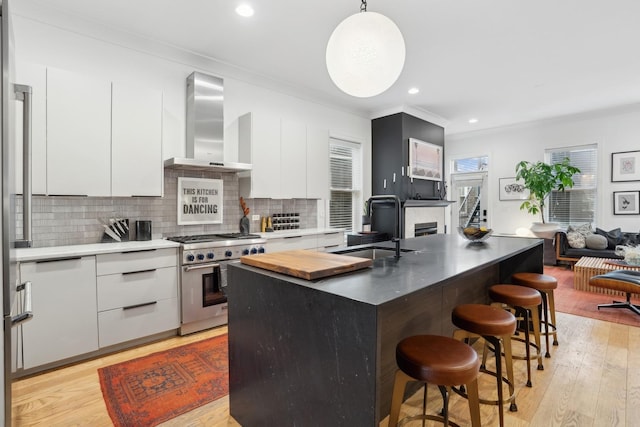 kitchen featuring high end range, pendant lighting, an island with sink, white cabinets, and wall chimney exhaust hood