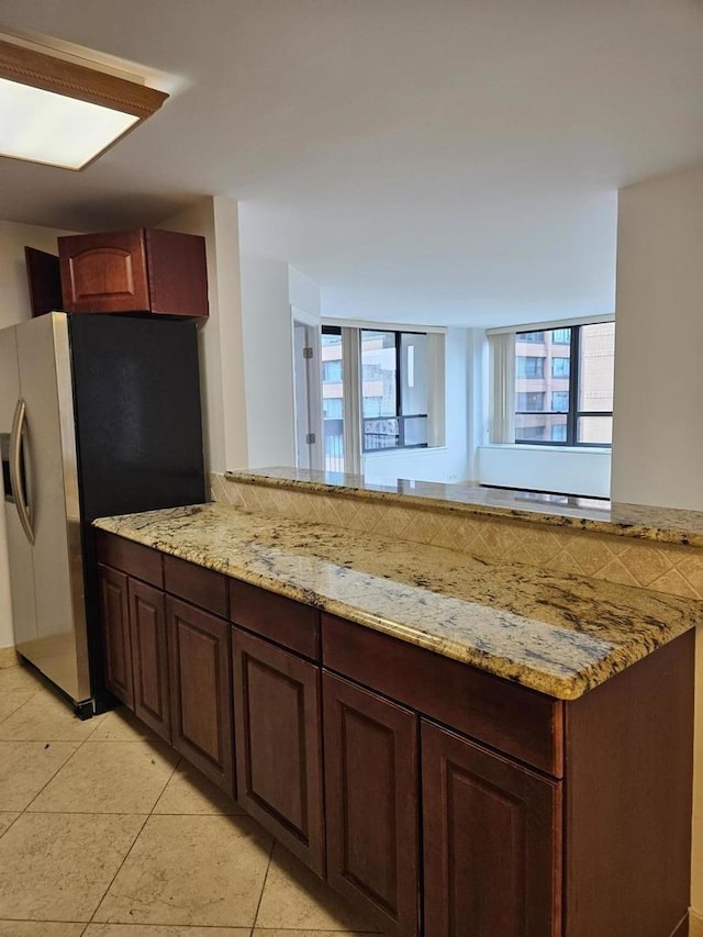 kitchen with kitchen peninsula, stainless steel refrigerator with ice dispenser, light stone countertops, light tile patterned floors, and dark brown cabinets