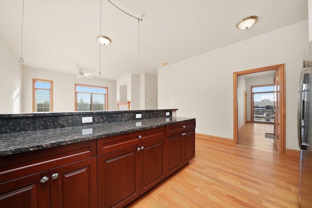 kitchen with ceiling fan, stainless steel refrigerator, light hardwood / wood-style flooring, hanging light fixtures, and dark stone counters