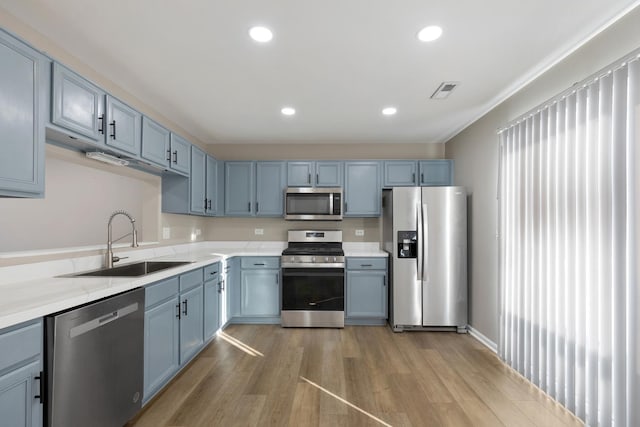 kitchen featuring appliances with stainless steel finishes, blue cabinetry, light wood-type flooring, light stone countertops, and sink