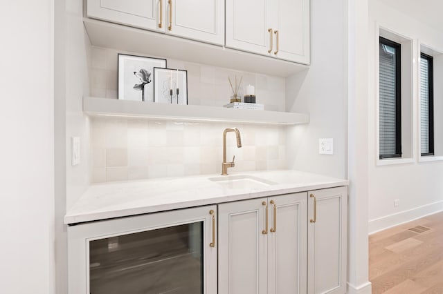 bar featuring wine cooler, decorative backsplash, sink, light wood-type flooring, and light stone countertops