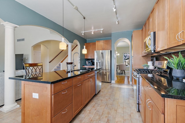 kitchen with sink, appliances with stainless steel finishes, an island with sink, decorative light fixtures, and ornate columns