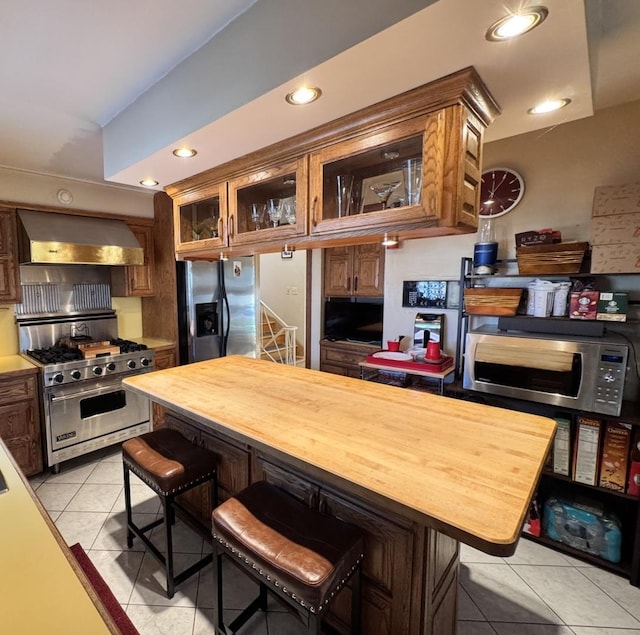 kitchen with a breakfast bar, butcher block countertops, light tile patterned flooring, stainless steel appliances, and wall chimney exhaust hood