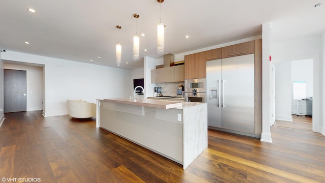 kitchen featuring backsplash, a center island with sink, hanging light fixtures, stainless steel built in fridge, and dark hardwood / wood-style flooring