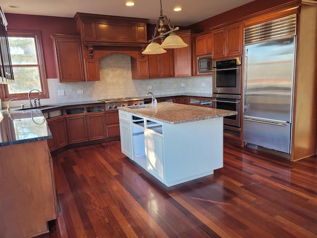 kitchen with a center island with sink, decorative light fixtures, dark wood-type flooring, light stone countertops, and built in appliances