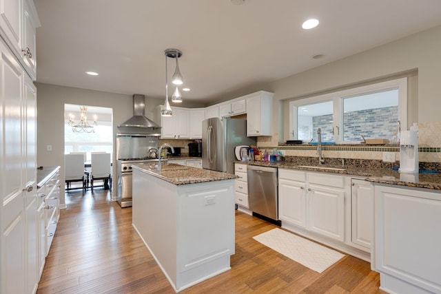 kitchen with an island with sink, white cabinetry, dark stone countertops, stainless steel appliances, and wall chimney range hood