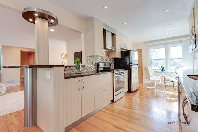 kitchen featuring appliances with stainless steel finishes, backsplash, dark stone counters, wall chimney exhaust hood, and light hardwood / wood-style flooring