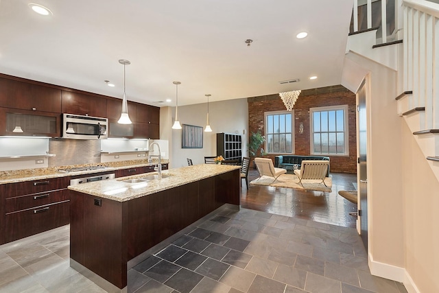 kitchen with sink, a kitchen island with sink, dark brown cabinets, stainless steel appliances, and light stone countertops