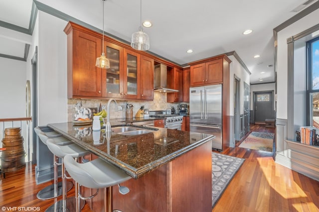 kitchen featuring sink, high end appliances, pendant lighting, dark stone counters, and wall chimney range hood