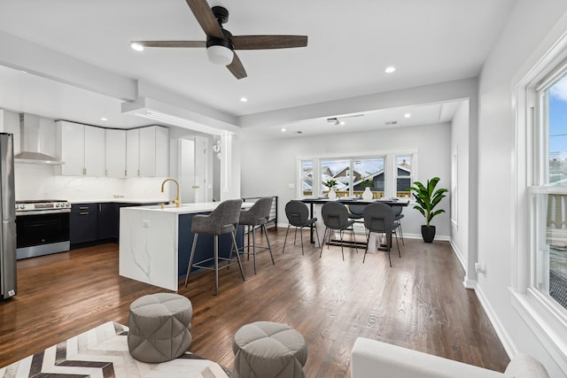 kitchen with wall chimney exhaust hood, a breakfast bar, appliances with stainless steel finishes, an island with sink, and white cabinets