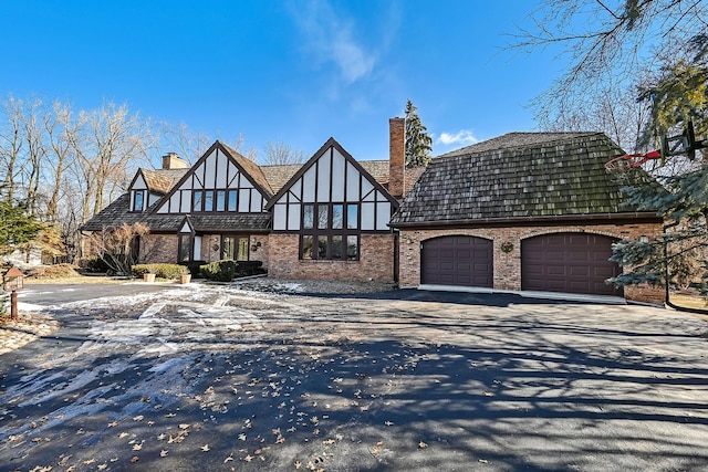 english style home with stucco siding, aphalt driveway, a garage, brick siding, and a chimney