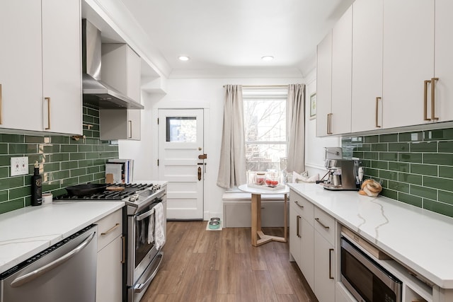 kitchen with wall chimney range hood, light hardwood / wood-style flooring, appliances with stainless steel finishes, backsplash, and white cabinets