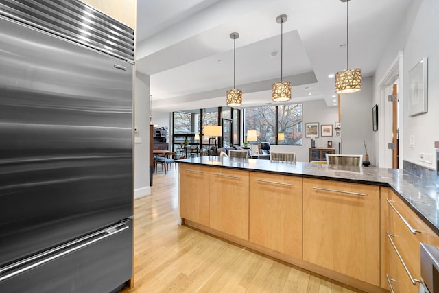 kitchen featuring decorative light fixtures, stainless steel built in refrigerator, dark stone counters, light brown cabinets, and light hardwood / wood-style flooring