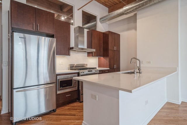 kitchen featuring wall chimney exhaust hood, sink, light hardwood / wood-style flooring, appliances with stainless steel finishes, and kitchen peninsula