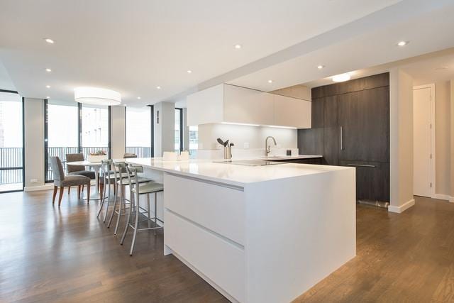 kitchen featuring white cabinets, dark brown cabinetry, dark wood-type flooring, a wall of windows, and a kitchen island with sink