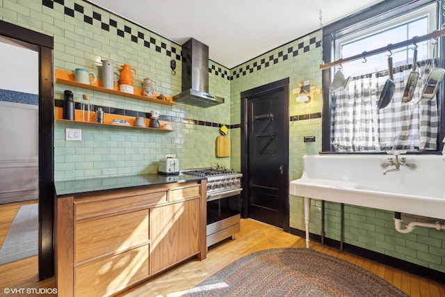 kitchen featuring light wood-style flooring, high end stainless steel range oven, decorative backsplash, wall chimney exhaust hood, and dark countertops