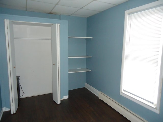 unfurnished bedroom with a closet, multiple windows, a paneled ceiling, dark wood-type flooring, and baseboard heating