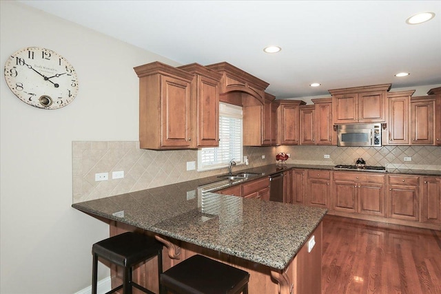 kitchen with brown cabinetry, a peninsula, dark stone counters, a sink, and stainless steel appliances