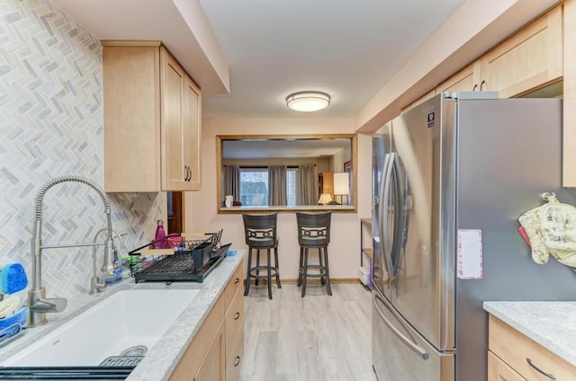 kitchen with light brown cabinetry, sink, backsplash, light stone counters, and stainless steel refrigerator