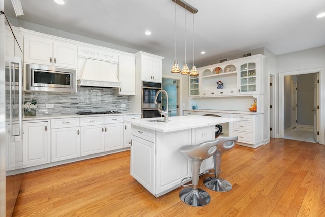 kitchen with pendant lighting, white cabinetry, stainless steel appliances, a center island with sink, and custom range hood