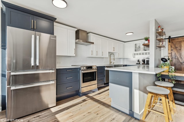 kitchen with a barn door, stainless steel appliances, a kitchen breakfast bar, wall chimney exhaust hood, and white cabinets