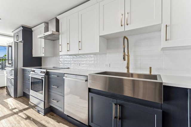 kitchen featuring white cabinets, appliances with stainless steel finishes, wall chimney range hood, light hardwood / wood-style floors, and sink