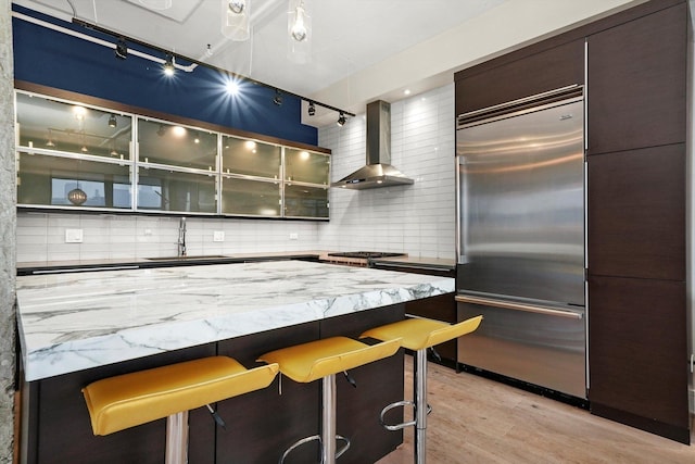 kitchen featuring stainless steel built in fridge, wall chimney range hood, light hardwood / wood-style flooring, a breakfast bar, and sink