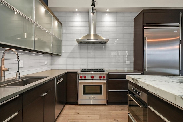 kitchen featuring wall chimney range hood, decorative backsplash, sink, high end appliances, and light wood-type flooring